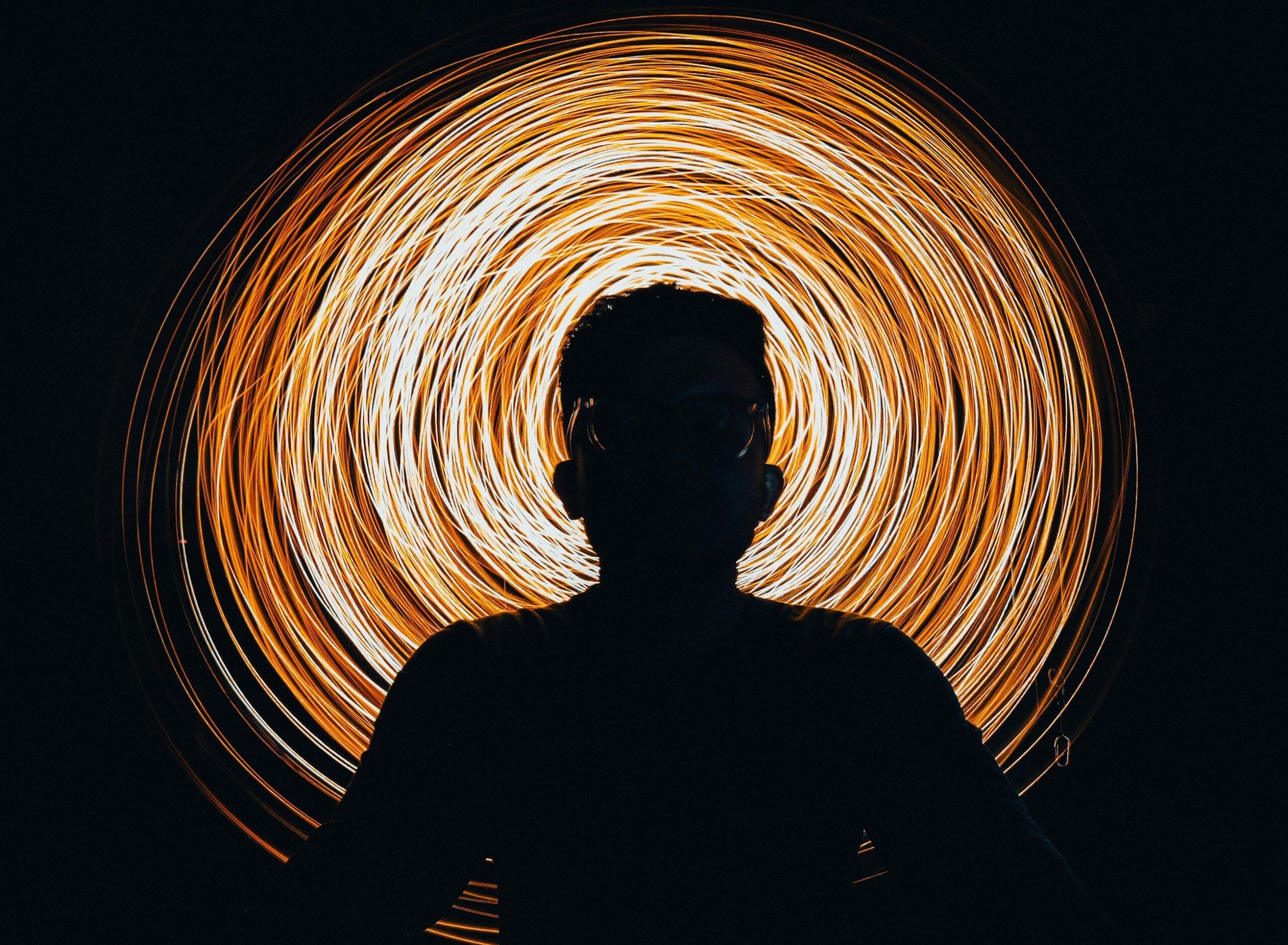 AI Summit Article image_part II Silhouette of a person surrounded by a circular swirl of golden light against a dark background.